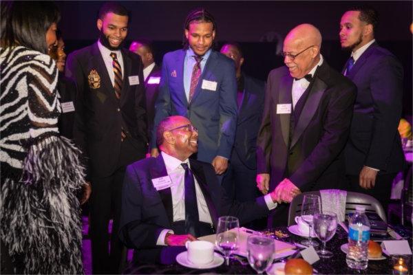 Bill Pickard seated at a table while five people crowd around him; man at left shakes Bill's hand